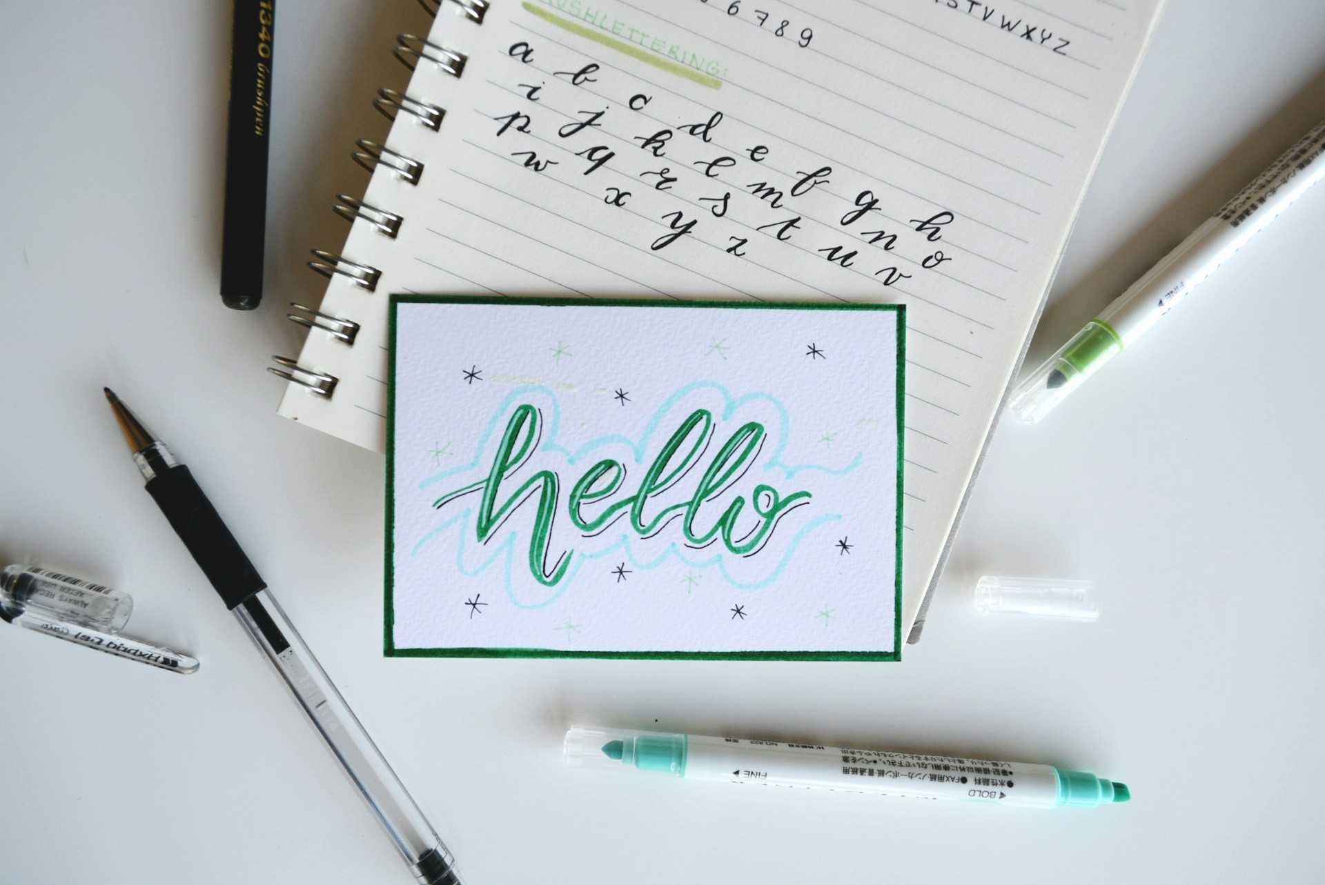 Table blanche avec un carnet, des stylos et une fiche avec écrit "Hello" en vert dessus
